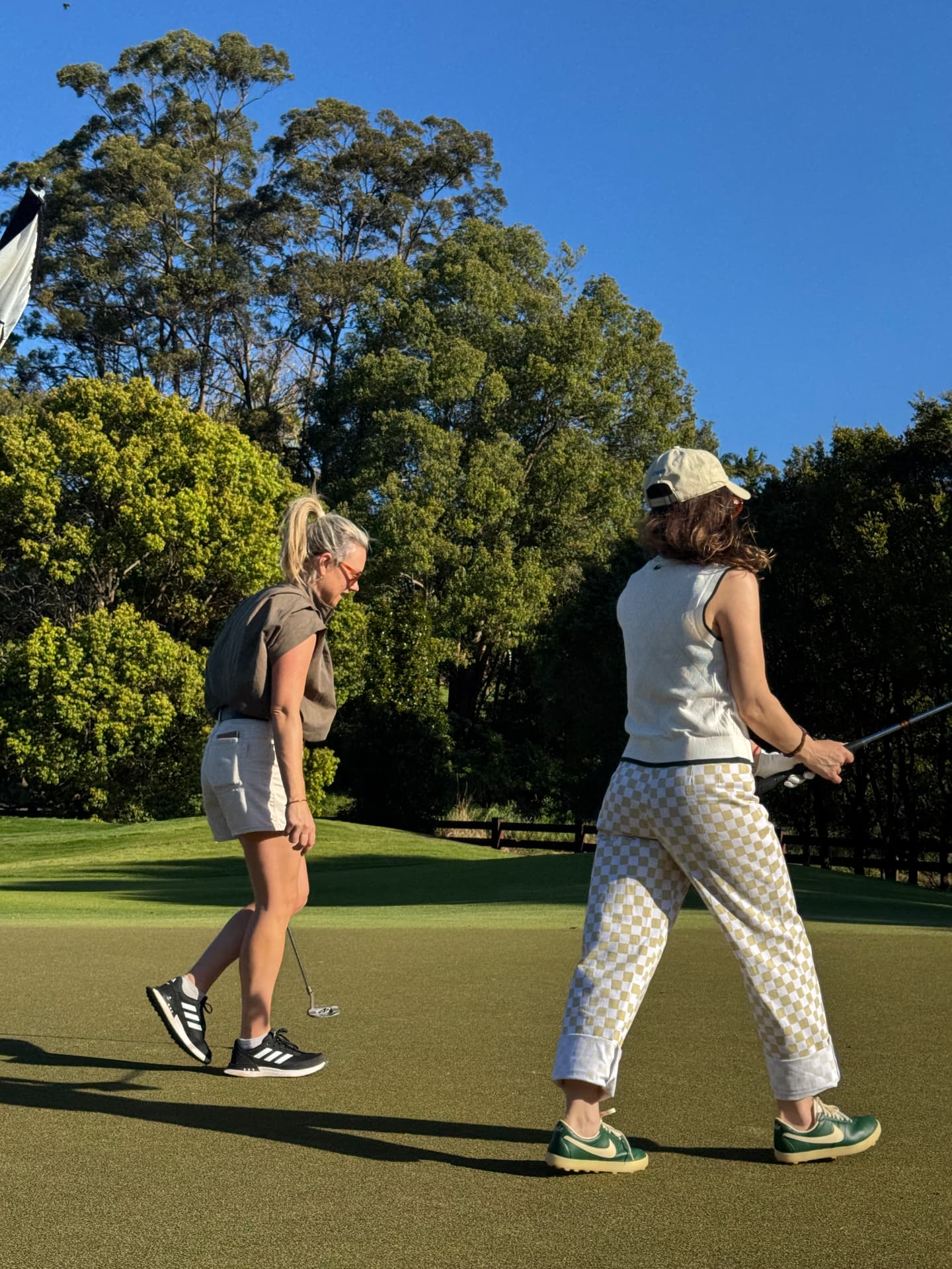 Two women walking across a sunny green with putters, one in checkered trousers